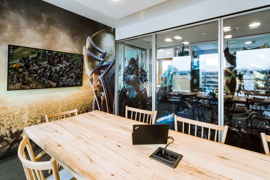 A meeting room with a wooden table, a large game artwork on the wall and a game on the screen